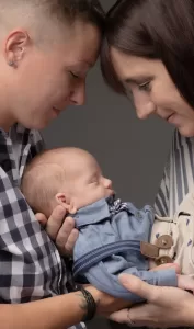 Professional Photographer Two adults gently touch foreheads while cradling a sleeping baby dressed in blue. The adults both look down at the baby lovingly, creating an intimate, tender portrait of family against a neutral background. www.maranonimagery.com