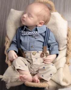 Professional Photographer A baby wearing a blue shirt, checkered bowtie, suspenders, and beige pants with small blue designs sleeps peacefully in a cozy basket lined with a cream-colored blanket, creating a portrait perfect for wedding photography inspiration. www.maranonimagery.com