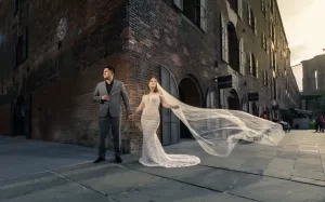 Professional Photographer A bride in a white lace gown with a long, flowing veil holds hands with a groom in a gray suit. They stand on a city street corner beside a brick building, sunlight streaming in from the right—perfect for timeless wedding photography. www.maranonimagery.com