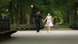 Professional Photographer A couple holding hands, smiling, and walking along a tree-lined park path with benches on either side. Perfect for capturing candid moments in wedding photography—the man wears black and the woman wears a white dress. www.maranonimagery.com