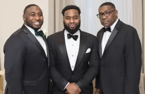 Professional Photographer Three men wearing black tuxedos with bow ties stand together indoors, posing and smiling for a photo. The background features light-colored curtains and furnishings, creating a classic setting perfect for wedding photography. www.maranonimagery.com