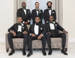 Professional Photographer Six men dressed in black tuxedos with bow ties and boutonnieres pose for a formal wedding photography portrait; three sit on a taupe couch while three stand behind them, all smiling or looking confidently at the photographer. www.maranonimagery.com