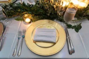 Professional Photographer A formal table setting with a gold charger plate, white folded napkin, silver cutlery, a water glass, champagne flute, candles, and green foliage centerpiece on a white tablecloth—perfect for wedding photography. www.maranonimagery.com