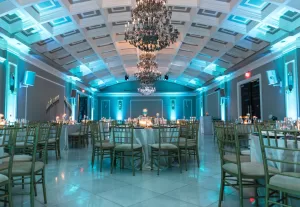 Professional Photographer A large, elegant banquet hall with round tables set for an event, gold chairs, candlelit centerpieces, crystal chandeliers, and blue accent lighting illuminating the high coffered ceiling—an exquisite venue perfect for memorable wedding photography. www.maranonimagery.com