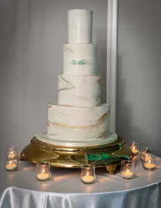 Professional Photographer A five-tiered marble-effect wedding cake on a gold stand, surrounded by small lit candles, set on a round table draped with a white tablecloth against a gray wall—perfectly captured by the photographer for timeless memories. www.maranonimagery.com