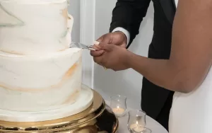 Professional Photographer A couple cuts a tiered wedding cake together, their hands joined on the knife. Perfect for wedding photography, the white cake with pastel accents sits on a gold stand, surrounded by small lit candles. www.maranonimagery.com