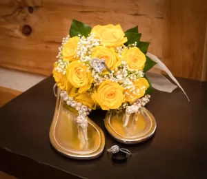 Professional Photographer Gold sandals adorned with rhinestones sit in front of a bouquet of yellow roses with babys breath. Two rings and a heart-shaped brooch rest among the flowers, creating a perfect scene for elegant wedding photography. www.maranonimagery.com