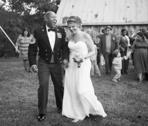 Professional Photographer A smiling couple, the man in a military dress uniform and the woman in a strapless wedding gown, walk hand in hand outdoors, surrounded by cheering guests at a rustic venue—a perfect moment for beautiful wedding photography. www.maranonimagery.com