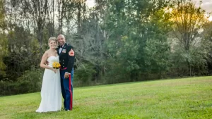 Professional Photographer A bride in a white dress and a groom in a decorated military uniform stand close together, smiling, on a grassy field with trees in the background—an unforgettable moment captured through expert wedding photography. www.maranonimagery.com