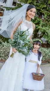 Professional Photographer A smiling bride in a white lace dress and veil holds a bouquet of greenery, standing beside a flower girl in a white dress and floral headband. This outdoor portrait captures their joy near lush green foliage. www.maranonimagery.com
