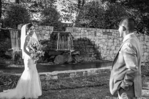 Professional Photographer A bride in a wedding dress and veil holds a bouquet, smiling at a man in a suit, as a photographer captures the moment near a stone wall with waterfalls and a small pond in an outdoor garden setting. www.maranonimagery.com
