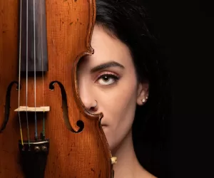 Professional Photographer A woman with dark hair holds a violin vertically, covering half of her face. Her uncovered eye looks directly at the camera, capturing an artistic portrait that highlights the creativity of a skilled photographer. The background is black. www.maranonimagery.com