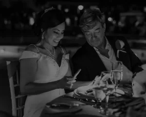 Professional Photographer A smiling woman in a white dress looks at her phone beside a man in a suit at a candlelit dinner table, captured by a wedding photographer. The scene, with glasses and plates before them, resembles portraits from an elegant celebration. www.maranonimagery.com