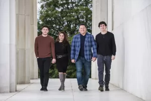 Professional Photographer Four people, two men and two young adults, walk together outdoors between marble columns. Smiling in casual clothes with trees behind them, the group looks ready for a perfect headshot or even candid wedding photography. www.maranonimagery.com