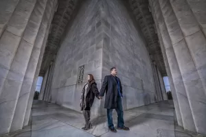 Professional Photographer A man and woman stand back-to-back holding hands at the corner of a large stone building with tall columns, looking in opposite directions. The dramatic, symmetrical scene captures the artistry of wedding photography. www.maranonimagery.com