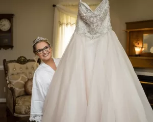 Professional Photographer A smiling woman in a white robe and tiara holds up a sparkling, strapless wedding gown in a warmly lit room with vintage decor, perfect for stunning portraits or headshots. www.maranonimagery.com