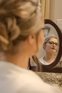 Professional Photographer A woman wearing glasses and a tiara looks thoughtfully at her reflection in a small oval mirror. Her hair is styled in an updo, embodying the elegance often captured in headshots or portraits, while standing by a granite countertop in a well-lit room. www.maranonimagery.com
