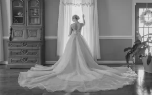 Professional Photographer A bride in a long, flowing gown stands facing a window with sheer curtains, her back to the camera, as the photographer captures this soft-lit portrait amid wooden furniture and a plant near the door. www.maranonimagery.com