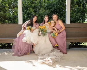 Professional Photographer Four women, including a bride in a white dress and three bridesmaids in matching mauve dresses, sit smiling on a bench under a gazebo, holding bouquets of sunflowers—an elegant moment of wedding photography. The bride's dress cascades onto the ground. www.maranonimagery.com