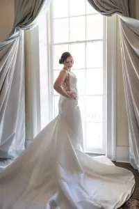 Professional Photographer A bride in a white, elegant wedding gown stands by a large window with flowing curtains, posing and smiling as natural light streams in behind her—capturing the artistry of wedding photography and timeless bridal portraits. www.maranonimagery.com