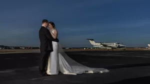Professional Photographer A bride and groom in wedding attire embrace on an airport tarmac, with a private jet and clear blue sky in the background—a striking moment perfect for unique portraits or romantic headshots. www.maranonimagery.com