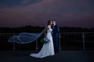 Professional Photographer A bride in a white gown and veil stands with a groom in a dark suit on a balcony at dusk. Perfect for wedding photography, the bride holds a bouquet as her veil flows behind her, framed by trees and a dark blue-pink sky. www.maranonimagery.com