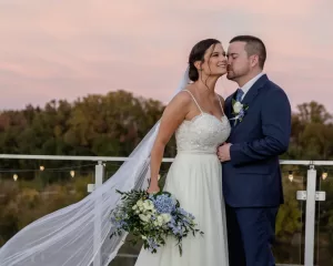 Professional Photographer A bride in a white dress and veil holds a bouquet of blue and white flowers while her groom, in a blue suit, kisses her forehead. Captured by a wedding photographer, they stand outdoors by a railing at sunset with trees in the background. www.maranonimagery.com