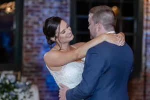 Professional Photographer A bride in a white dress smiles and embraces a groom in a blue suit as they dance together—an intimate moment captured through elegant wedding photography, with a blurred brick wall and windows in the background. www.maranonimagery.com