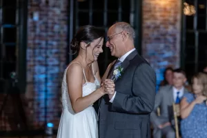 Professional Photographer A bride in a white dress smiles and holds hands with an older man in a suit, dancing together at a wedding reception. A photographer captures this heartfelt portrait amid brick walls and seated guests in the background. www.maranonimagery.com