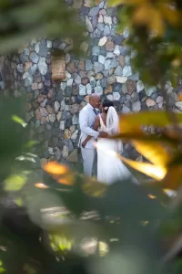 Professional Photographer A bride and groom embrace in front of a stone wall, framed by blurred greenery. Captured by a talented wedding photographer, the couple appears joyful in this intimate outdoor portrait, dressed elegantly for their special day. www.maranonimagery.com