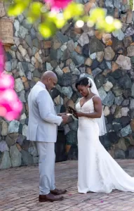 Professional Photographer A bride and groom stand facing each other, reading vows outdoors. Captured by a photographer, the bride wears a white gown and veil, the groom a light gray suit. They are against a stone wall with pink flowers blurred in the foreground. www.maranonimagery.com