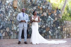 Professional Photographer A bride in a white dress and a groom in a light gray suit stand smiling and laughing together outdoors in front of a multicolored stone wall, perfectly captured by their photographer for beautiful portraits. www.maranonimagery.com