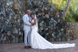 Professional Photographer A bride in a white wedding dress and a groom in a light suit embrace and smile in front of a stone wall outdoors. Captured by a skilled photographer, the scene feels like one of their most cherished portraits, with the bride’s train spread out behind her. www.maranonimagery.com