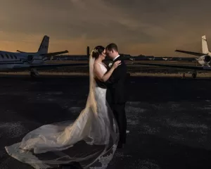 Professional Photographer A photographer captures a bride and groom embracing on an airport tarmac at sunset, standing between two private jets. The bride’s white dress flows behind her as the groom, in a black tuxedo, holds her close in this unforgettable moment. www.maranonimagery.com