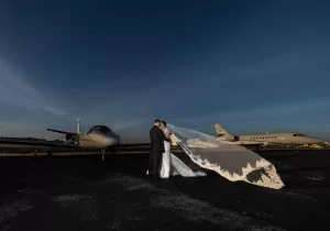 Professional Photographer A bride and groom embrace on an airport tarmac at sunset, her long veil flowing behind them. Captured by a talented photographer, two private jets sit in the background under a clear, darkening sky. www.maranonimagery.com
