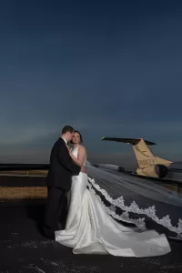Professional Photographer A photographer captures a bride and groom embracing on an airport runway at dusk, her lace veil flowing and a private jet in the background—a stunning moment in wedding photography. www.maranonimagery.com