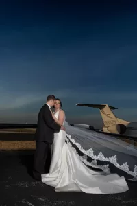 Professional Photographer A bride in a white gown and veil stands with a groom in a dark suit on an airport tarmac at dusk, as their photographer captures stunning portraits with her veil flowing in the wind and a private jet in the background. www.maranonimagery.com