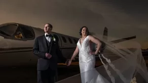 Professional Photographer A bride and groom in formal wedding attire stand hand in hand before a private jet. Captured by a skilled wedding photographer, the bride’s long veil flows in the wind as they gaze into the distance under a dramatic sky. www.maranonimagery.com