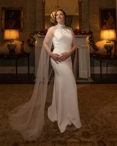 Professional Photographer A woman in a long, elegant white gown with sheer cape sleeves stands smiling in a warmly lit, classic living room—an ideal setting for stunning wedding photography by an experienced photographer. A fireplace and glowing lamps complete the scene. www.maranonimagery.com