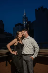 Professional Photographer A couple poses on a rooftop at night, captured by a skilled photographer; the woman wears a black dress and the man a white shirt with gray pants. City skyscrapers, including a lit-up tower, fill the background. www.maranonimagery.com