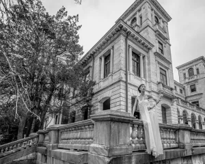 Professional Photographer A woman in a flowing dress stands on the steps of a large, historic building with classical architecture, captured in black and white by a skilled photographer, framed by stone railings and leafless trees. www.maranonimagery.com