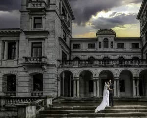Professional Photographer A bride and groom stand close together on the steps of a grand, historic building with arches and columns at sunset, as a photographer captures their portrait beneath a dramatic cloudy sky. www.maranonimagery.com