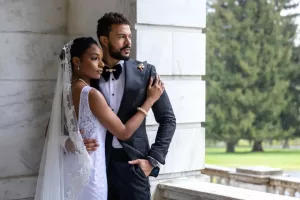 Professional Photographer A bride in a white dress and veil embraces a groom in a black tuxedo as they stand together by a marble column, looking thoughtfully into the distance. Lush greenery surrounds the couple, captured beautifully by a skilled photographer. www.maranonimagery.com