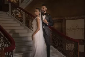 Professional Photographer A bride in a white gown and a groom in a black tuxedo stand on an elegant staircase with ornate railings, looking serious—a captivating moment of wedding photography in a grand, dimly lit setting. www.maranonimagery.com