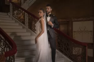 Professional Photographer A bride in a white gown and a groom in a black tuxedo stand together on an elegant staircase with ornate railings, as their photographer captures this formal wedding photo. www.maranonimagery.com