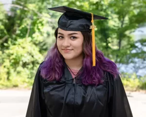 Professional Photographer A young woman with purple hair, wearing a black graduation cap and gown with a gold 2024 tassel, stands outdoors in front of green trees, smiling at the camera—an ideal portrait moment for any photographer. www.maranonimagery.com