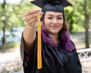 Professional Photographer A graduate in a black cap and gown holds out a gold 2021 tassel, smiling proudly with blurred greenery in the background—a perfect moment for graduation portraits. www.maranonimagery.com
