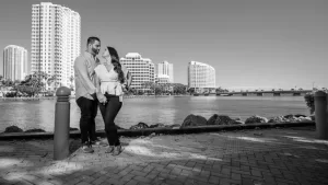 Professional Photographer A couple stands close together by a waterfront, smiling at each other. Tall buildings and a bridge are visible across the water under a clear sky. Captured by a talented photographer, the photo is beautifully composed in black and white. www.maranonimagery.com