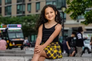 Professional Photographer A young girl with long curly hair sits on a stone bench outdoors, smiling at the camera. She wears a black tank top and a yellow plaid skirt. Perfect for portraits, blurred people, trees, and buildings appear in the background. www.maranonimagery.com