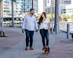 Professional Photographer A smiling couple walks hand in hand along a waterfront path lined with palm trees, while the woman holds a leash for their small brown dog. Perfect for pet photography with tall buildings and water in the background. www.maranonimagery.com
